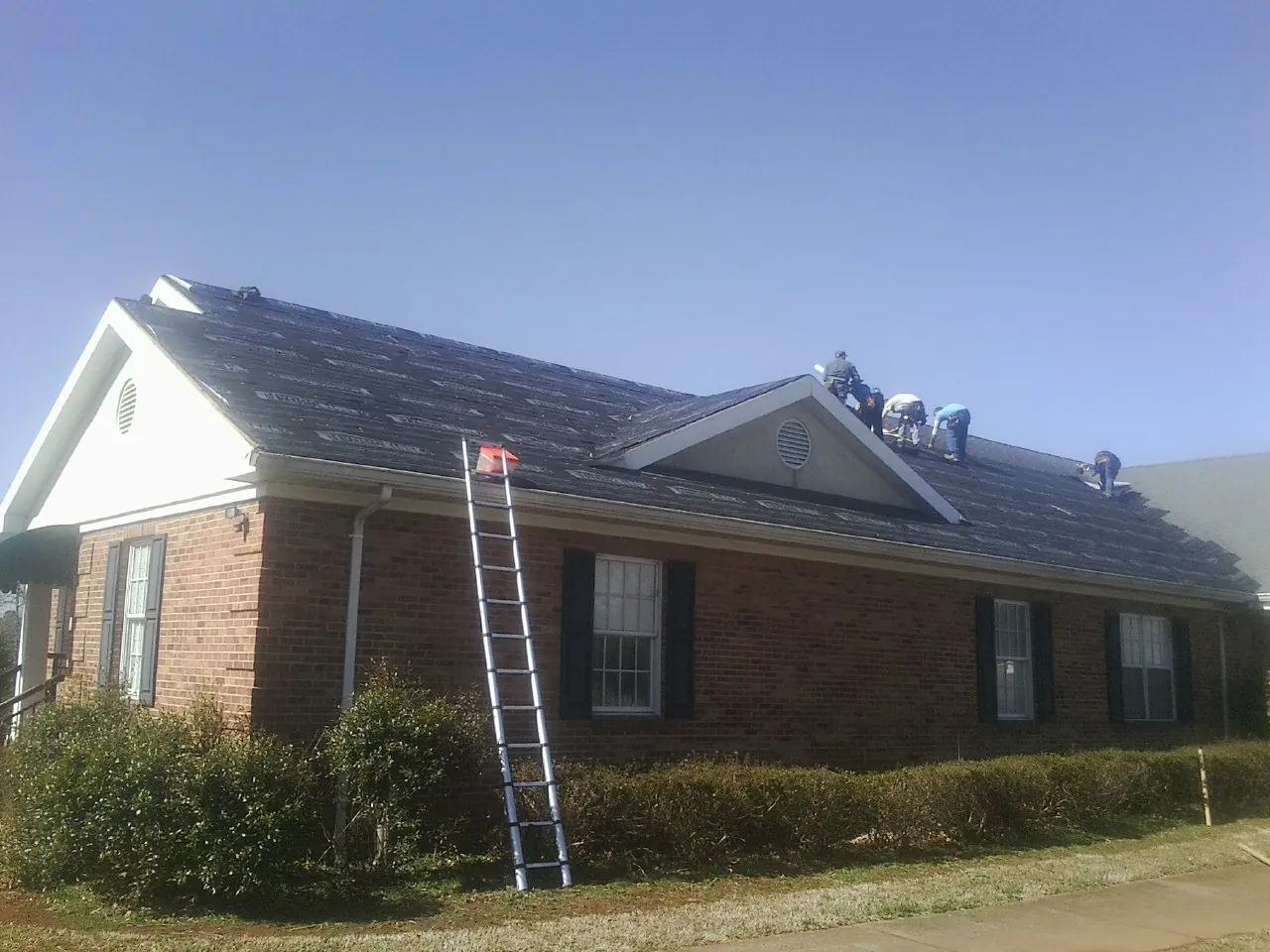 Crew tearing shingles off a brick ranch roof with a ladder set — Emergency Roof Repair work by our crew in Ocean Gate