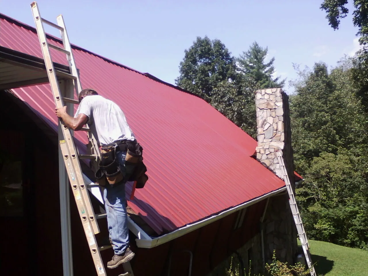 Roofer on a ladder beside a red metal roof and stone chimney — Soffit & Fascia Repair by Roofing Brothers in Ocean Gate